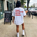 A person standing on a sidewalk facing a store, wearing a white oversized t-shirt with the text 'PARKS BALLENTINE HAITES HEMMES' printed on the back.