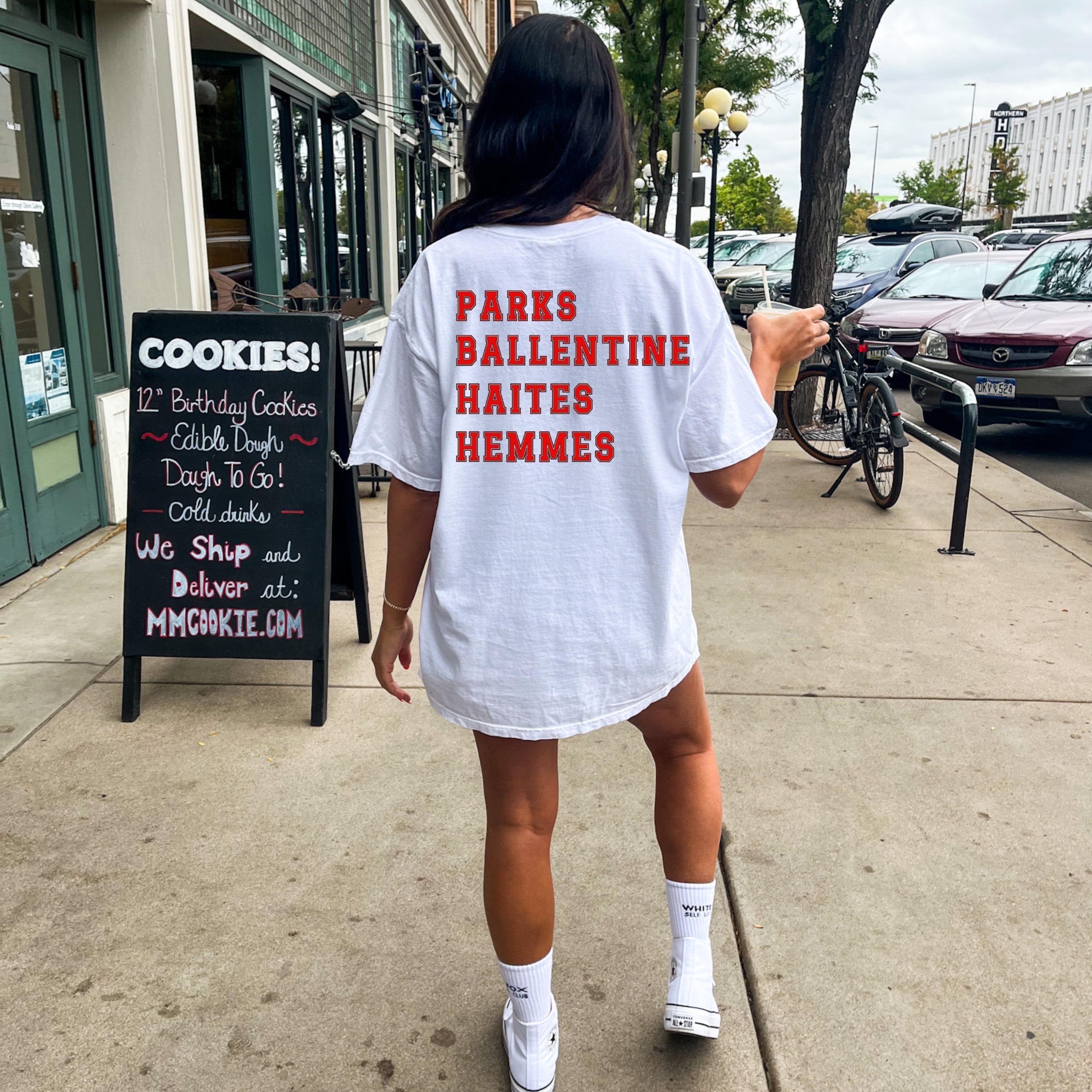 A person standing on a sidewalk facing a store, wearing a white oversized t-shirt with the text 'PARKS BALLENTINE HAITES HEMMES' printed on the back.