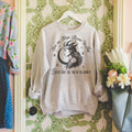 A gray sweatshirt with a graphic print inspired by the book 'A Fate Inked in Blood', displayed on a hanger against a wall with floral wallpaper.