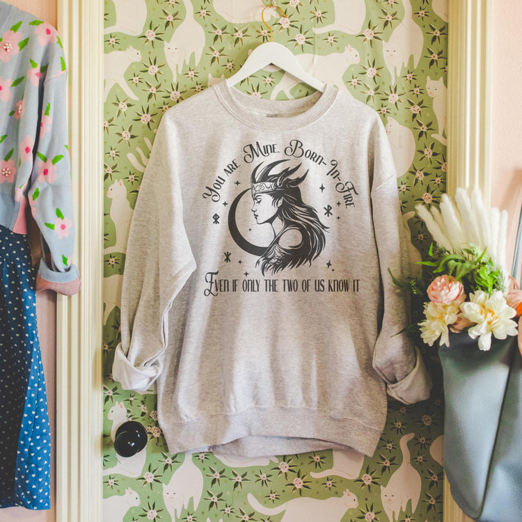 A gray sweatshirt with a graphic print inspired by the book 'A Fate Inked in Blood', displayed on a hanger against a wall with floral wallpaper.