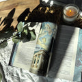 Fantasy-inspired travel mug with celestial illustrations and handwritten quote, resting on an open book with sunlight, eucalyptus, and a lit candle.