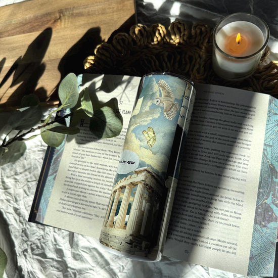 Fantasy-inspired travel mug with celestial illustrations and handwritten quote, resting on an open book with sunlight, eucalyptus, and a lit candle.