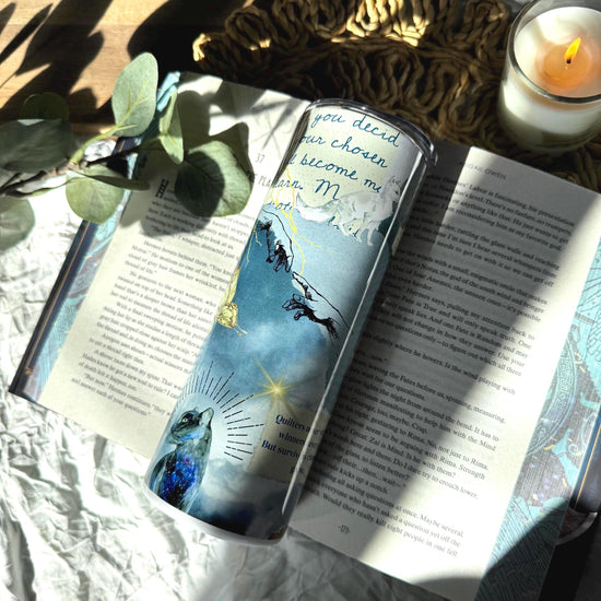 Fantasy-inspired 20 oz travel mug with divine hand illustration and celestial art, styled on an open book with candle and greenery in sunlight.