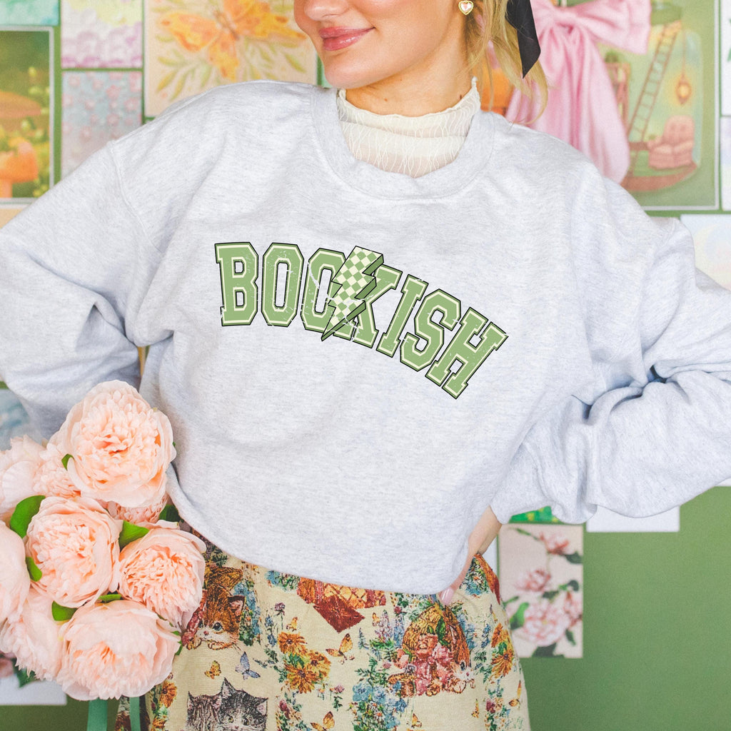 A person wearing a gray sweatshirt with the word 'BOOKISH' printed on the front in green letters, accompanied by a floral design in the background and a bundle of flowers to the side.