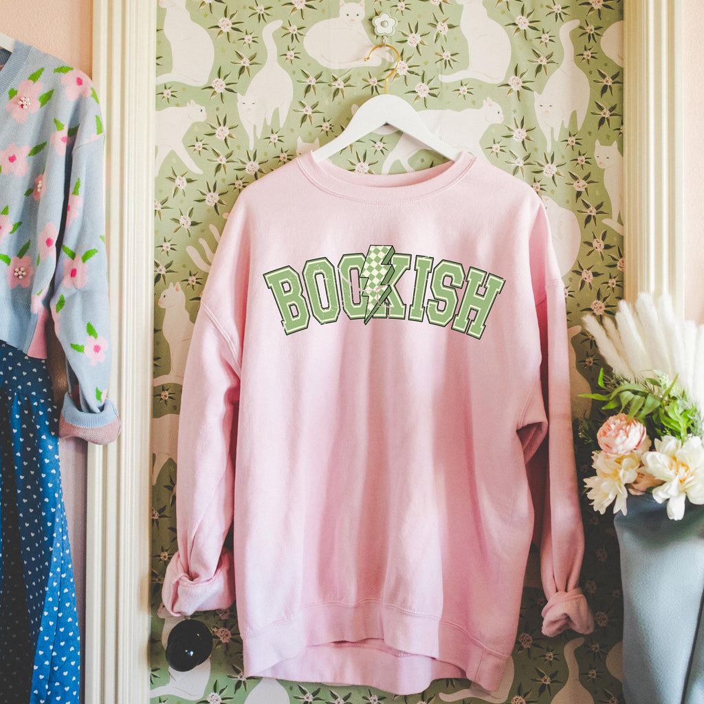 Pink Bookish Bolt sweatshirt with whimsical lightning/book graphic, styled with books and cozy reading props.