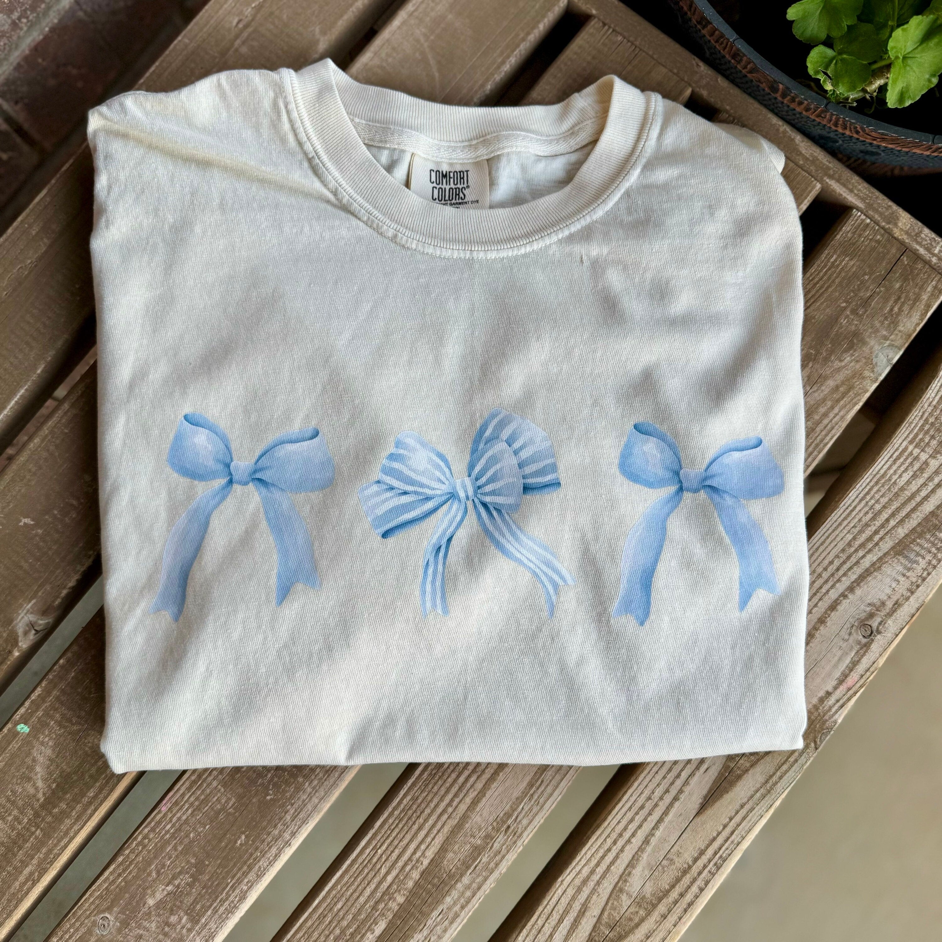 A top-down view of the ivory Coquette Bows Cropped Tee laid flat. The image shows the full front of the garment, highlighting the soft ivory cotton and the three blue bow graphics.