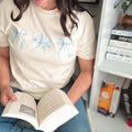An ivory cropped t-shirt featuring light blue bow graphics, styled in a quiet reading corner. The scene includes an open book and a white bookshelf, capturing a still moment in a personal library
