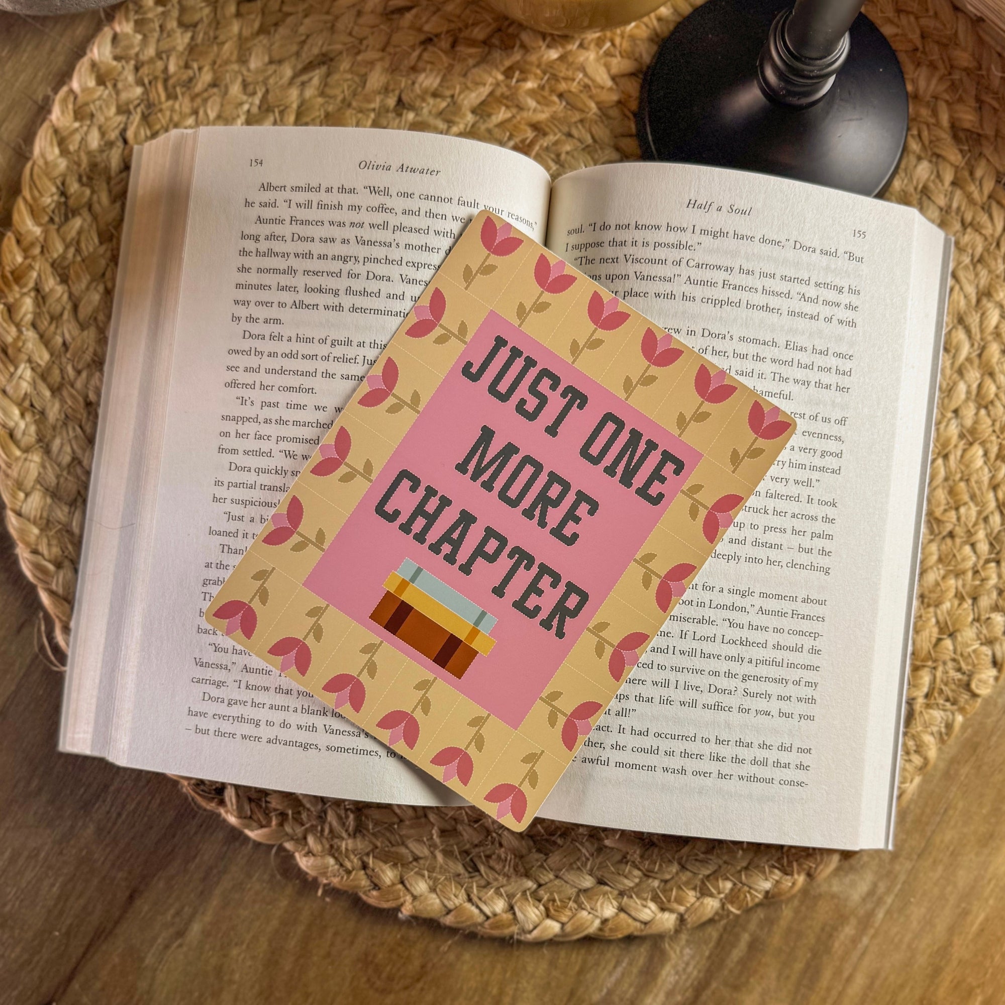 A lifestyle flat lay featuring a 'Just One More Chapter' e-reader insert with a floral border, placed on the open pages of an old book. The warm lighting and wooden surface emphasize the cozy, nostalgic feel of the reader's accessory.