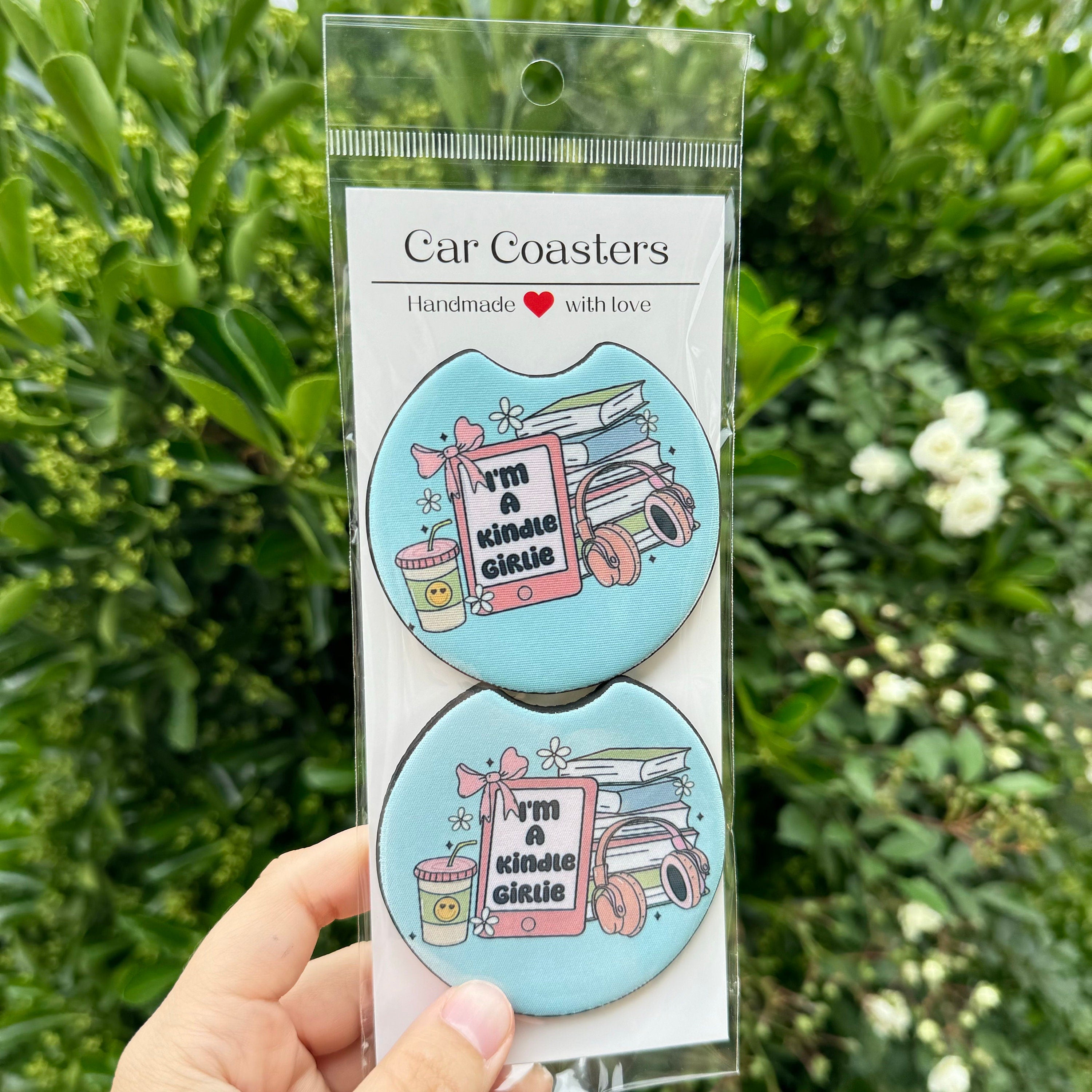 A 2-pack of blue Kindle Girlie car coasters by Dex Avenue, featuring bookish icons and a reading quote, styled in a clear package against a garden background