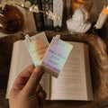 Hand holding two library-card style holographic stickers featuring the quote "Well-Read Even on My Worst Days" in a Dark Academia library setting.