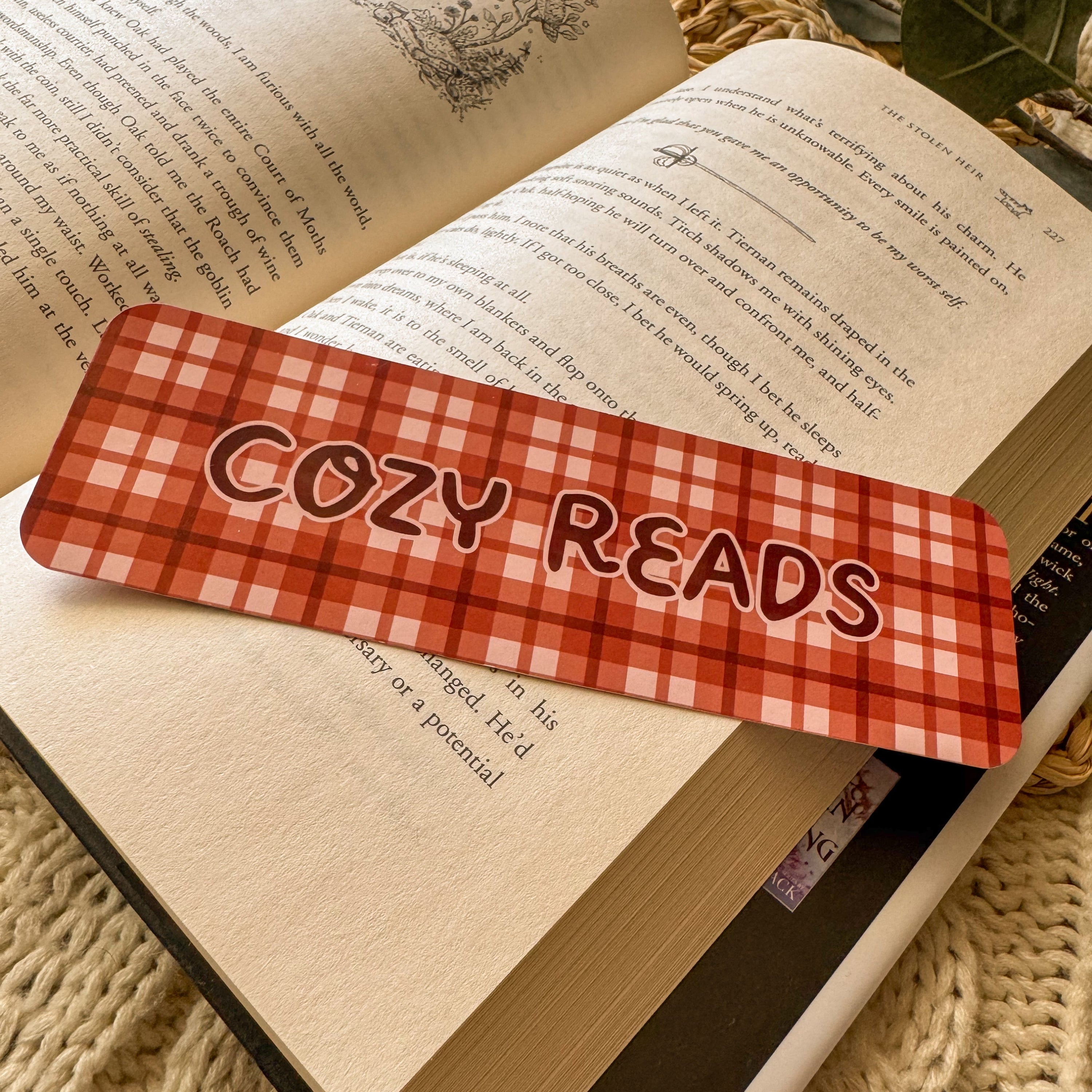 A close-up of a maroon and white checkered glossy bookmark labeled 'Cozy Reads,' styled on an open book to highlight the smooth, substantial paper quality.