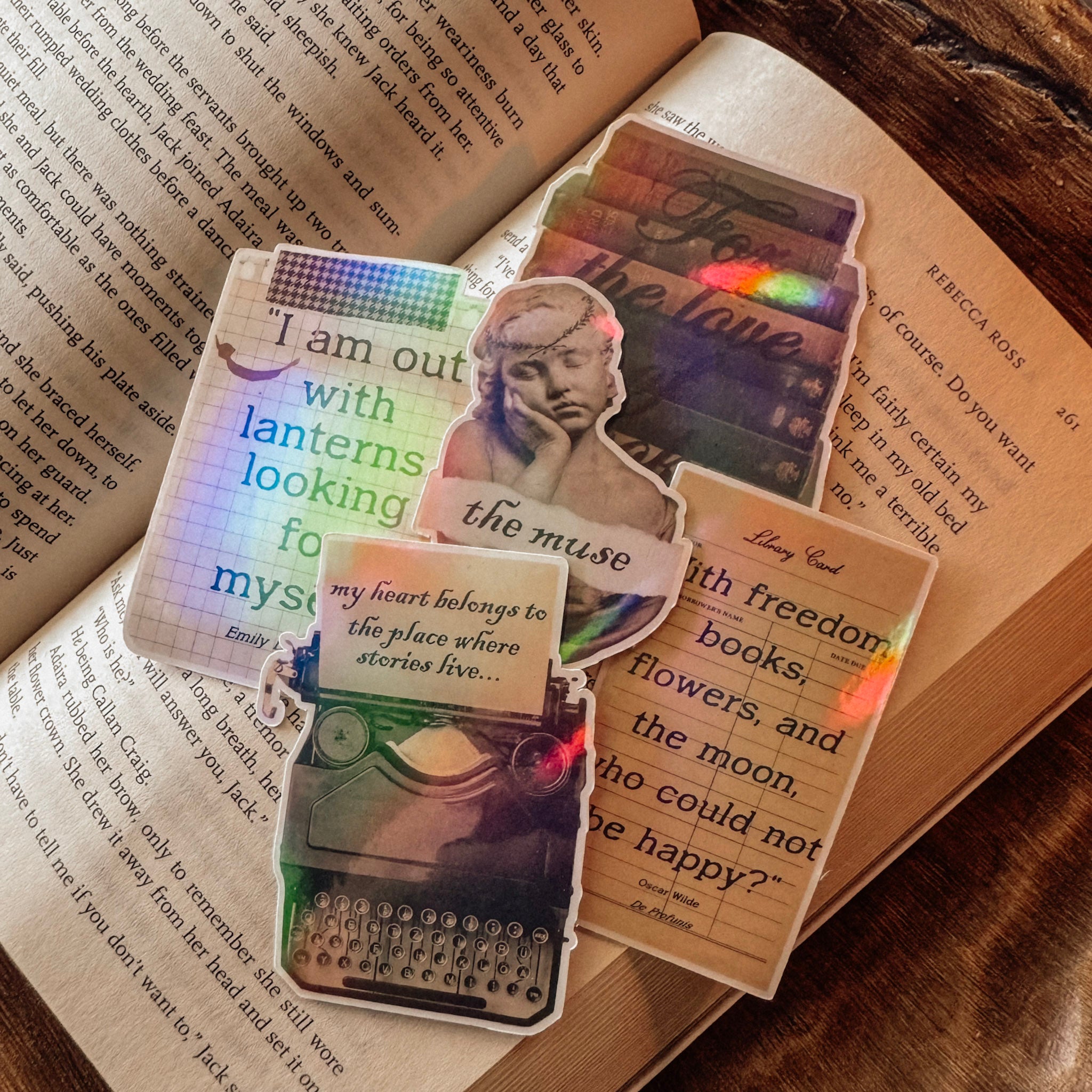 A flat lay of the Muse and Manuscript collection featuring five holographic stickers: Oscar Wilde quote, Emily Dickinson lantern, a vintage typewriter, a classical marble bust, and a stack of antique books. The stickers show a vibrant rainbow shimmer against a dark academia background.