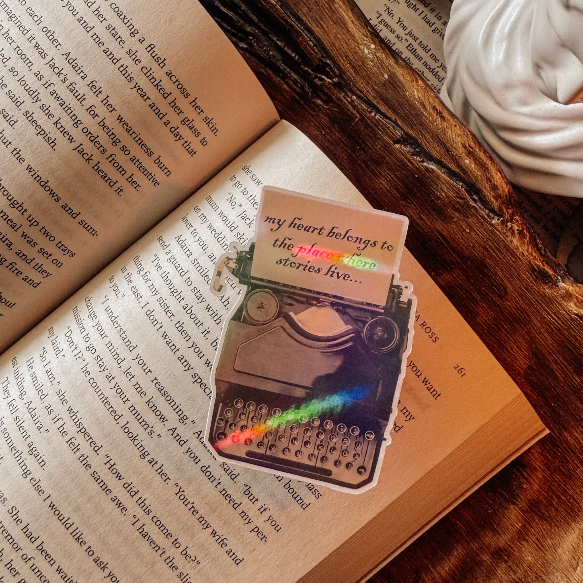 Holographic sticker of a vintage typewriter with the quote "My heart belongs to the place where stories live." Styled in a light academia aesthetic on a vintage book page next to a marble statue bust and a lit candle with a prismatic rainbow shimmer.