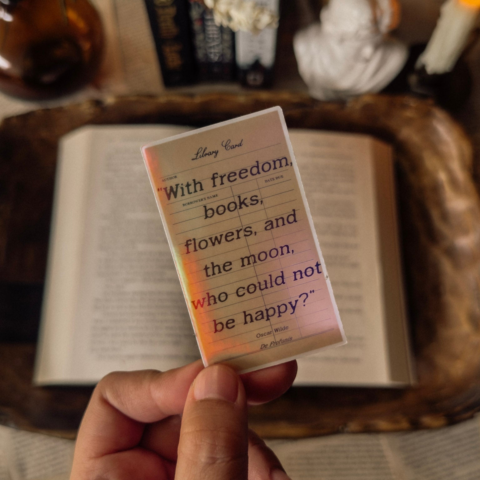 Close-up of a holographic Oscar Wilde quote sticker "With freedom, books, flowers, and the moon, who could not be happy?" The sticker displays a vibrant rainbow prismatic effect, styled on an antique book page with a marble bust in a dark academia aesthetic.
