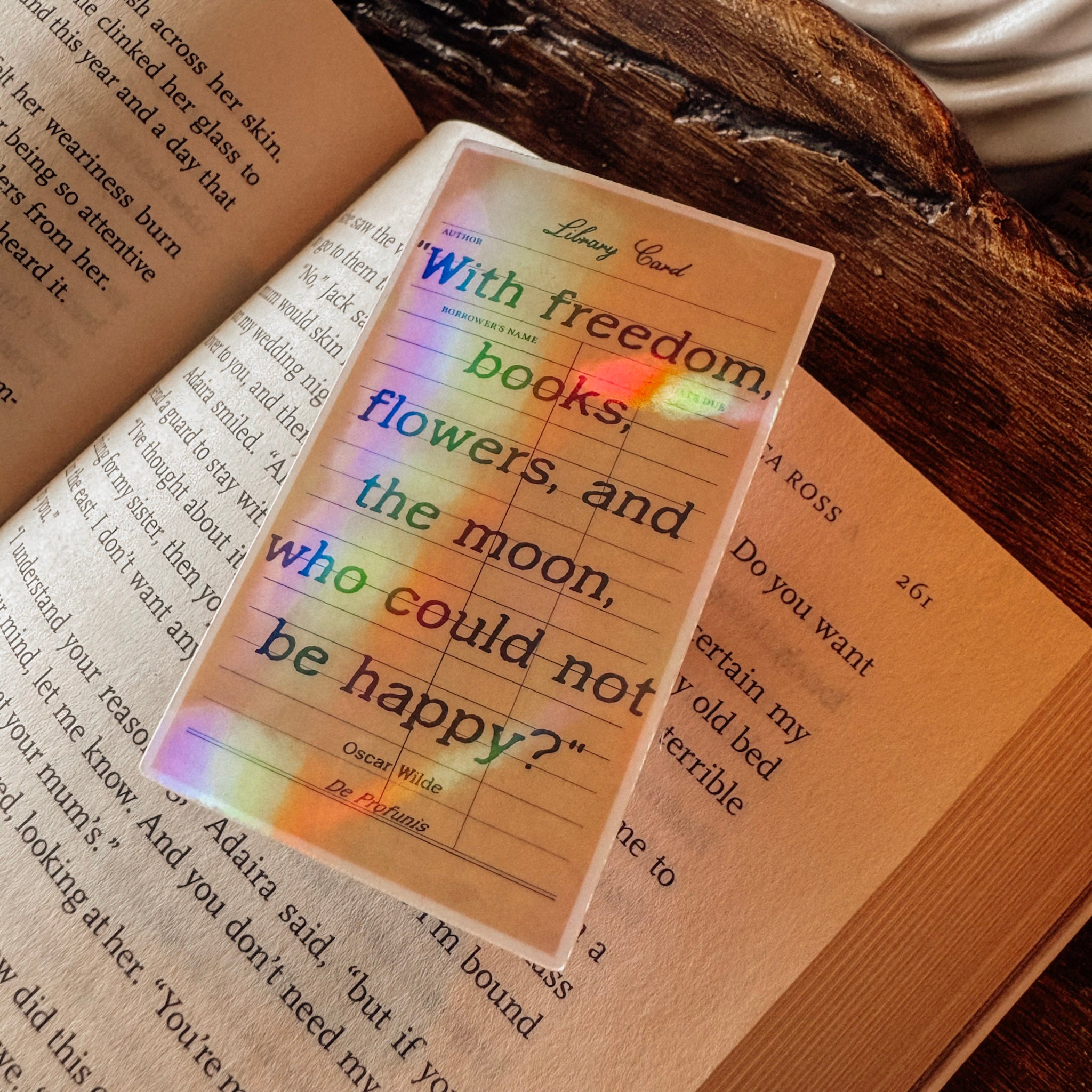 Holographic Oscar Wilde quote sticker featuring the text "With freedom, books, flowers, and the moon, who could not be happy?" Styled on a vintage book with a classical marble bust and a prismatic rainbow shimmer in a light academia setting.