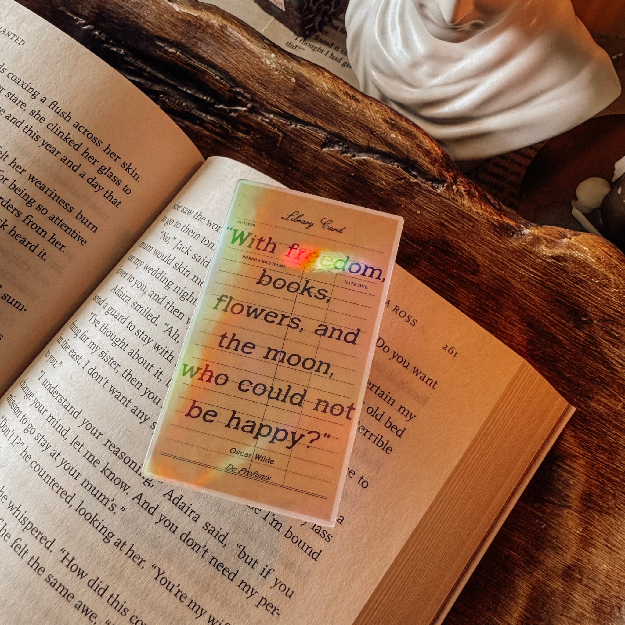 Handheld shot of a 3-inch holographic Oscar Wilde quote sticker "With freedom, books, flowers, and the moon, who could not be happy?" The image shows the prismatic rainbow effect and the sticker's size against a dark academia backdrop of vintage books and a marble bust.