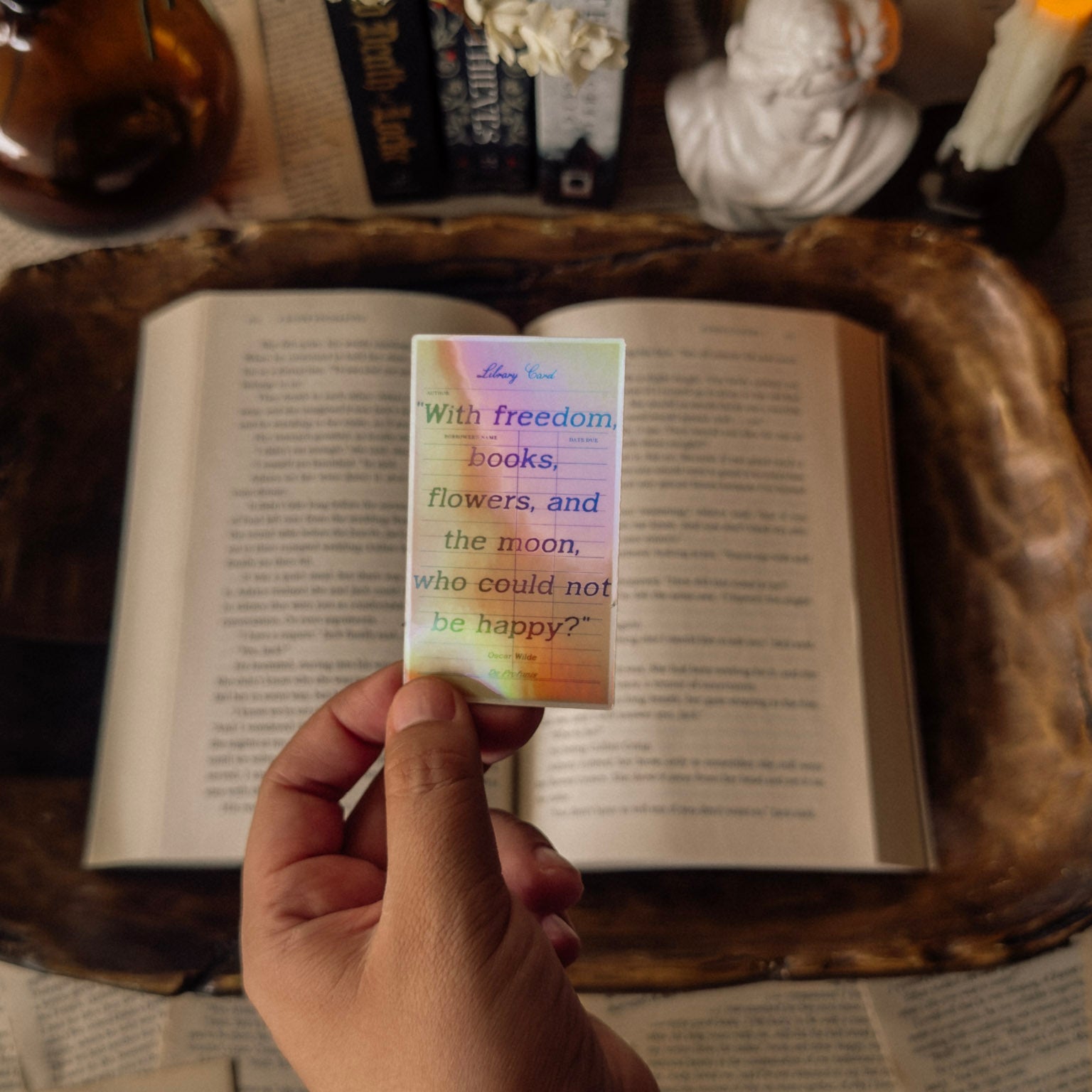 Holographic Oscar Wilde sticker with the quote "With freedom, books, flowers, and the moon, who could not be happy?" Styled in a dark academia aesthetic on a vintage book next to a marble statue and a lit candle with a prismatic rainbow shimmer.