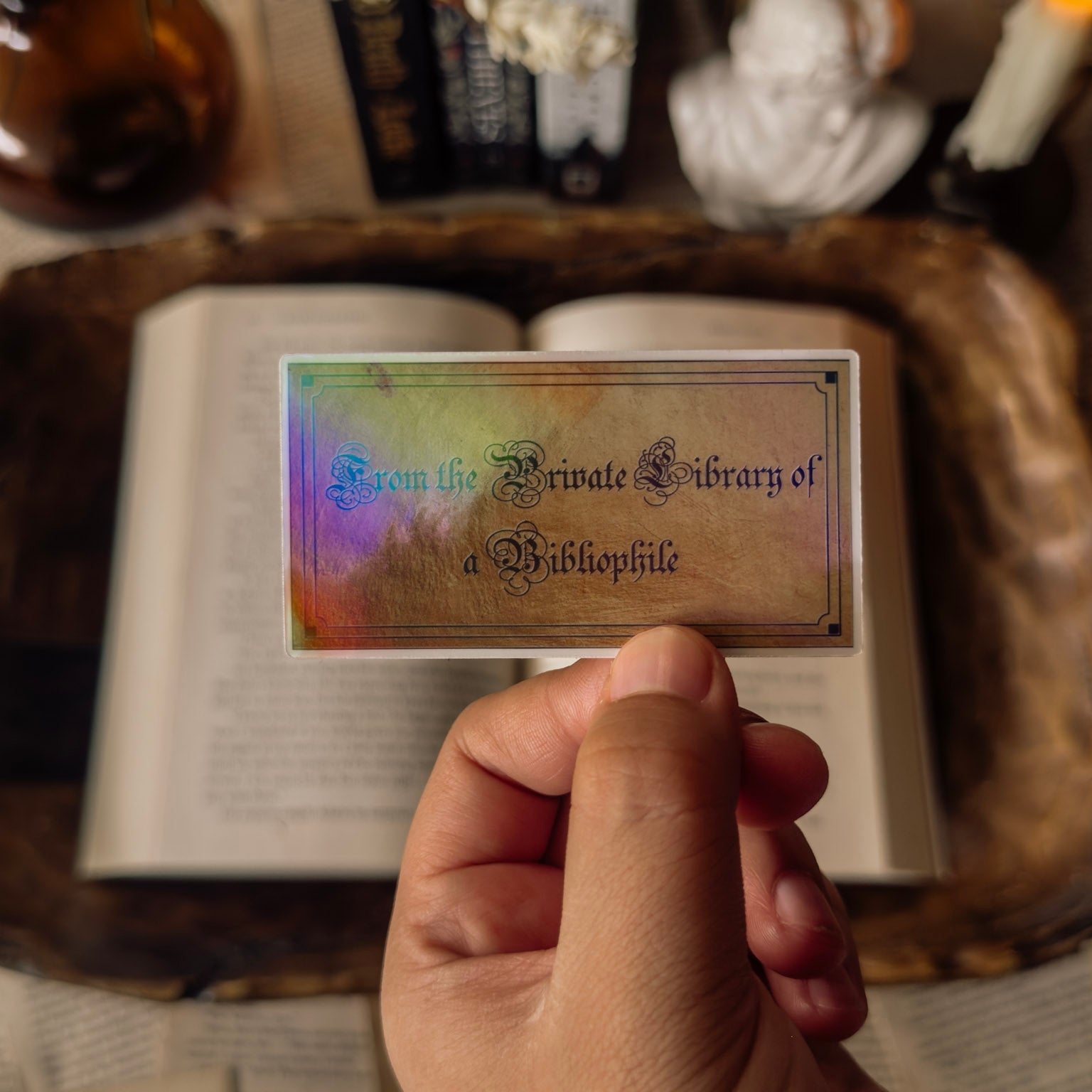 Close up of a hand holding a holographic 'From the Private Library of a Bibliophile' sticker against a moody dark academia background with an open book and candles.