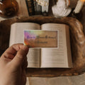 A hand holding a rectangular holographic sticker that says 'From the Private Library of a Bibliophile' over an open book with a dark academia aesthetic background