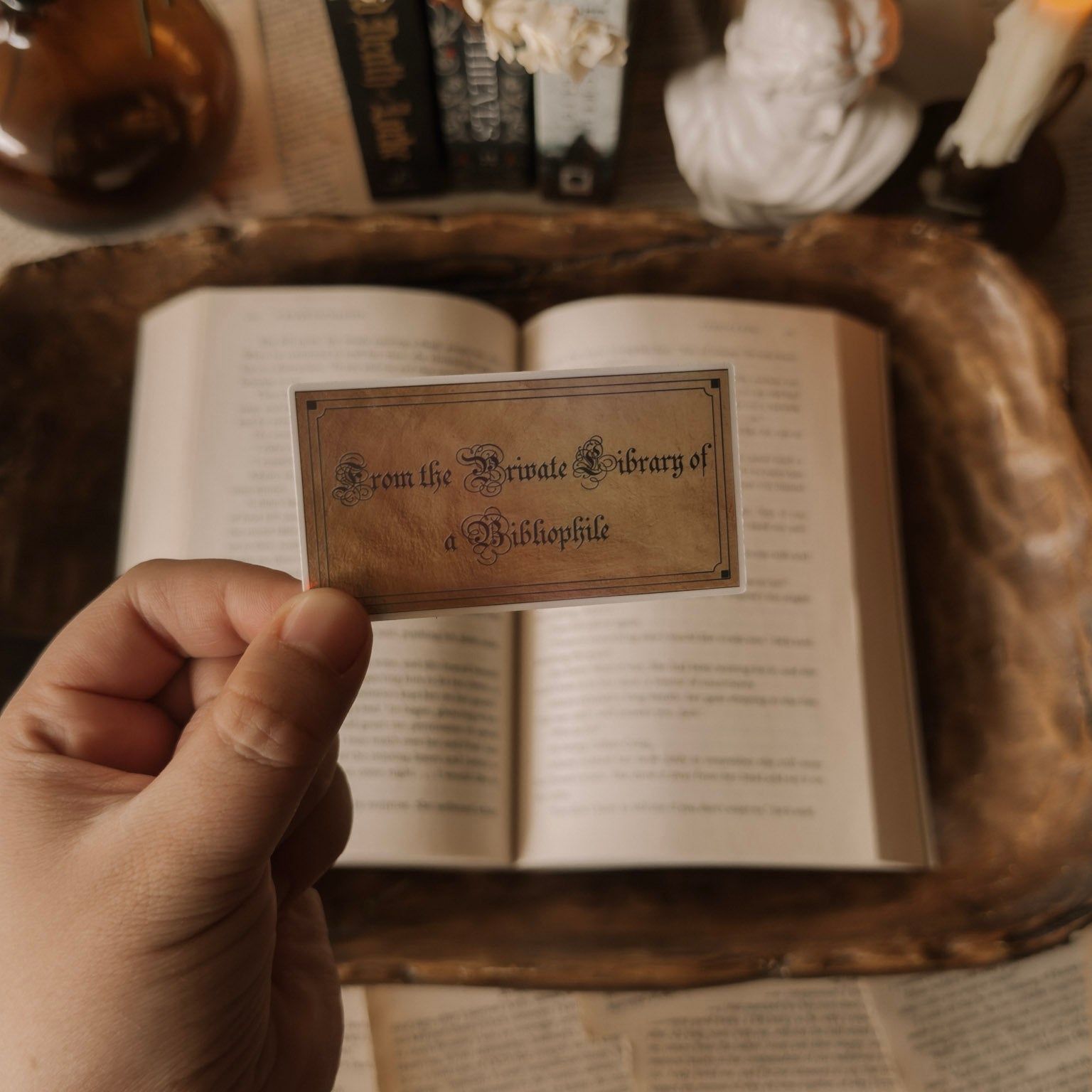 A hand holding a holographic 'Private Library of a Bibliophile' sticker over an open vintage book, surrounded by a dark academia setting with candles and old volumes.