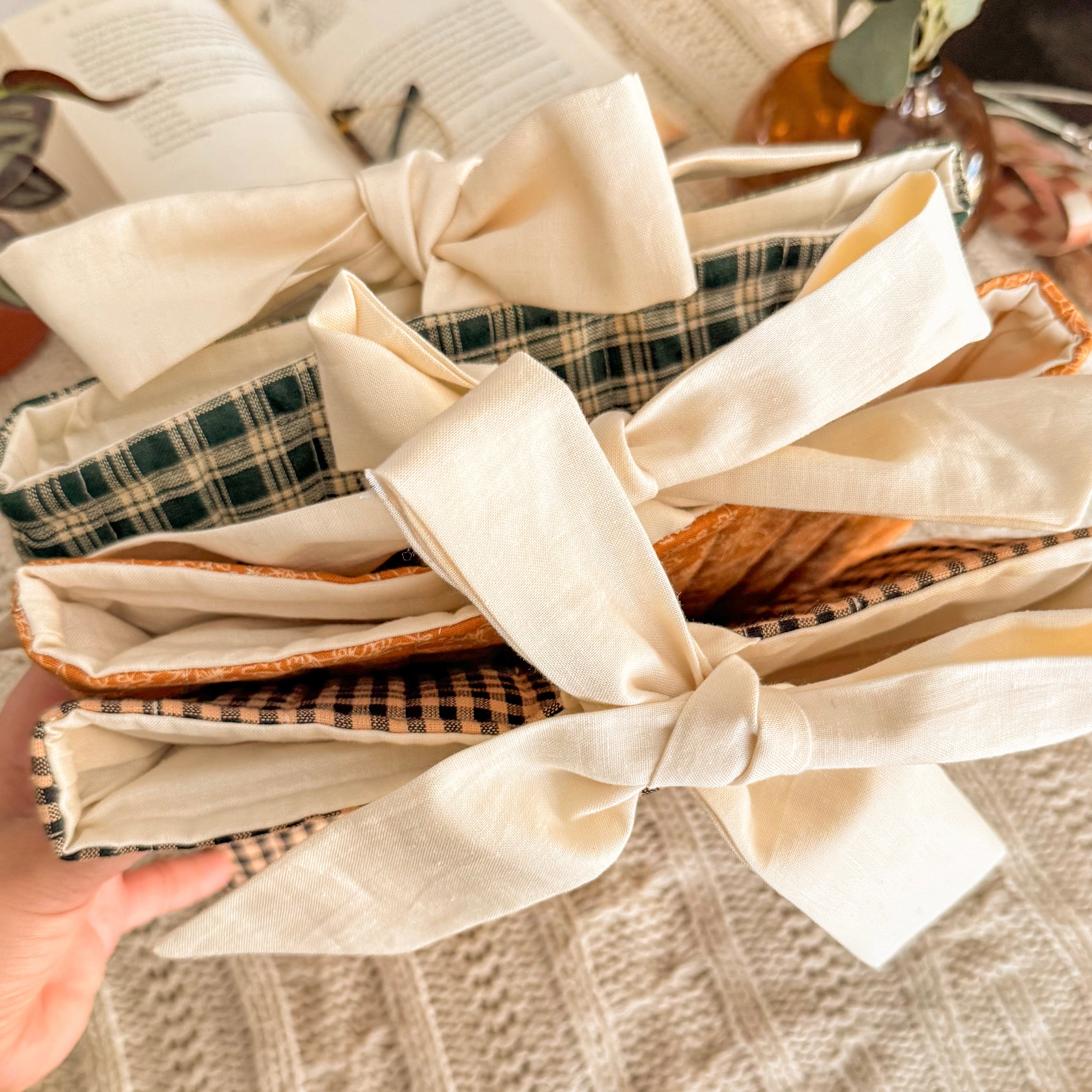 A stack of three handmade Quilted Book Sleeves in green gingham, orange floral, and brown plaid patterns, featuring large cream cotton bow closures in a cozy reading setting.
