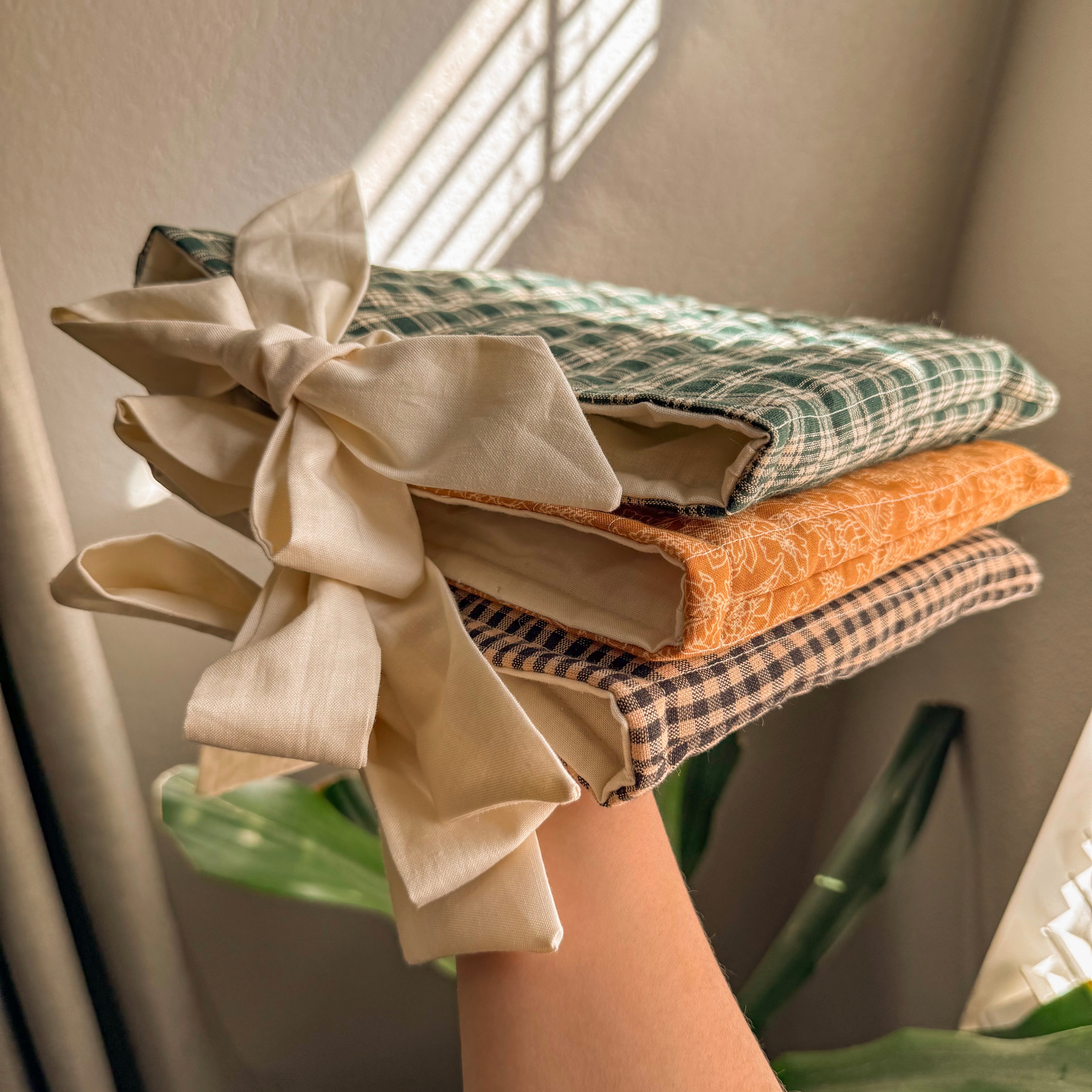 A hand holding a stack of three handmade Quilted Book Sleeves in green gingham, orange floral, and brown plaid patterns, featuring cream fabric bows in soft natural sunlight.
