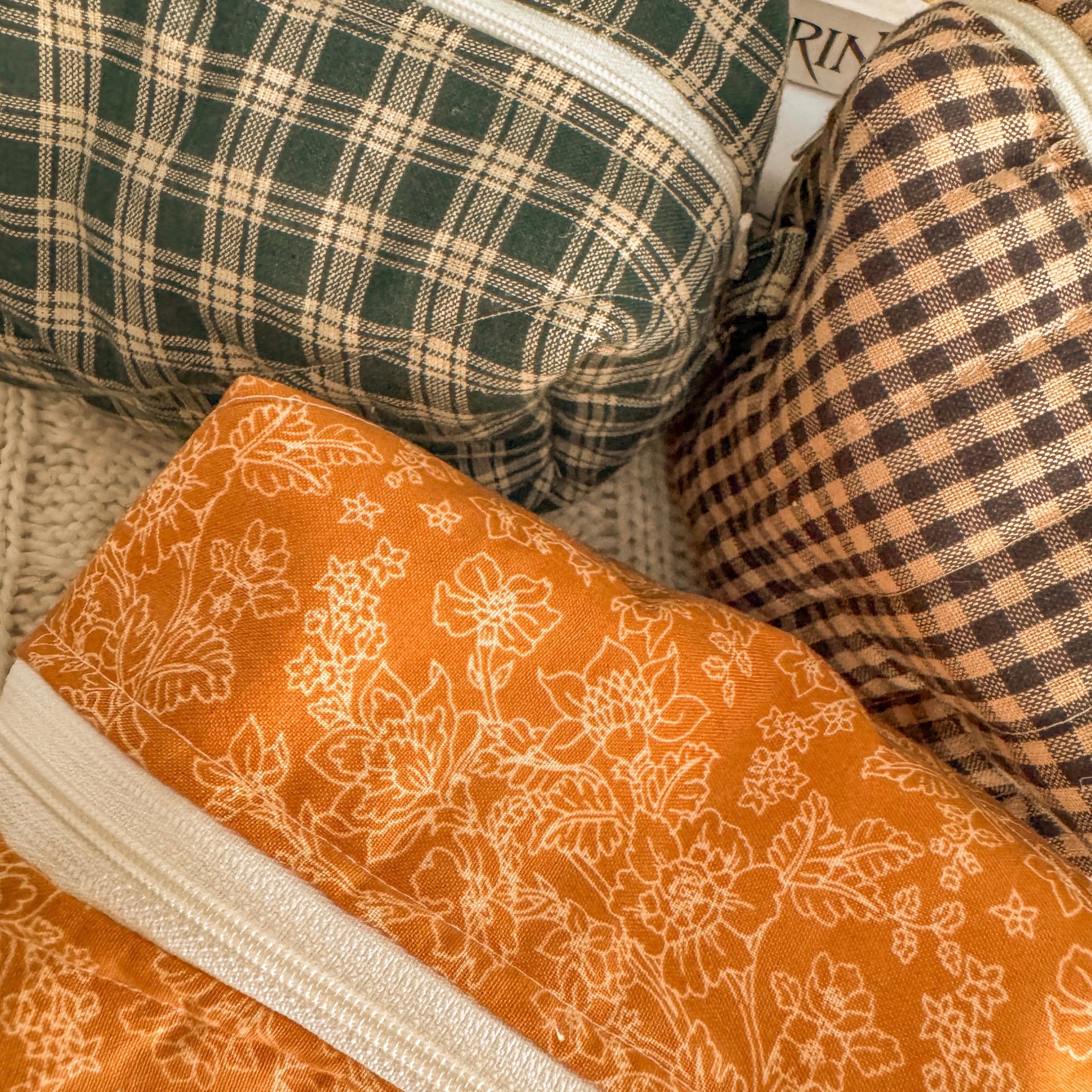A close-up detail shot of three quilted fabric pouches, highlighting the intricate textures of the orange floral, green plaid, and brown checkered patterns. The warm lighting emphasizes the handcrafted quality of the materials.
