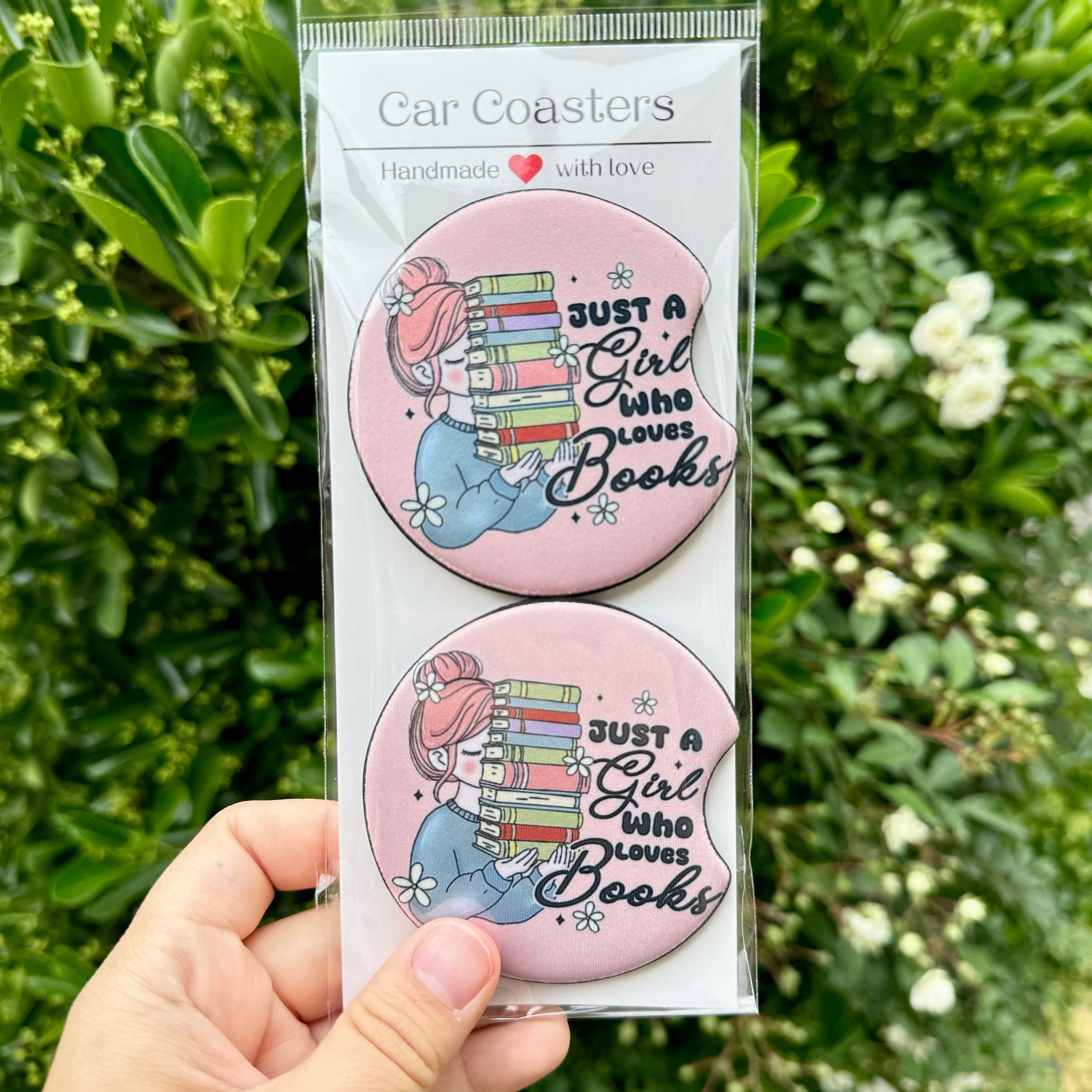 A 2-pack of Reader Girl car coasters by Dex Avenue, featuring a pink design with a book stack and broom illustration, packaged and ready for a car cup holder