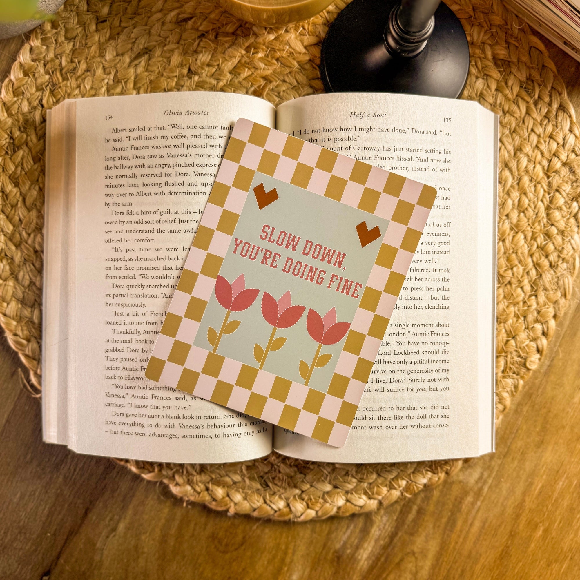 A lifestyle photo of a 'Slow Down You're Doing Fine' quilted Kindle insert resting on the pages of an open vintage book. Styled on a woven coaster with a warm glass of coffee in the background for a cozy, nostalgic reading aesthetic