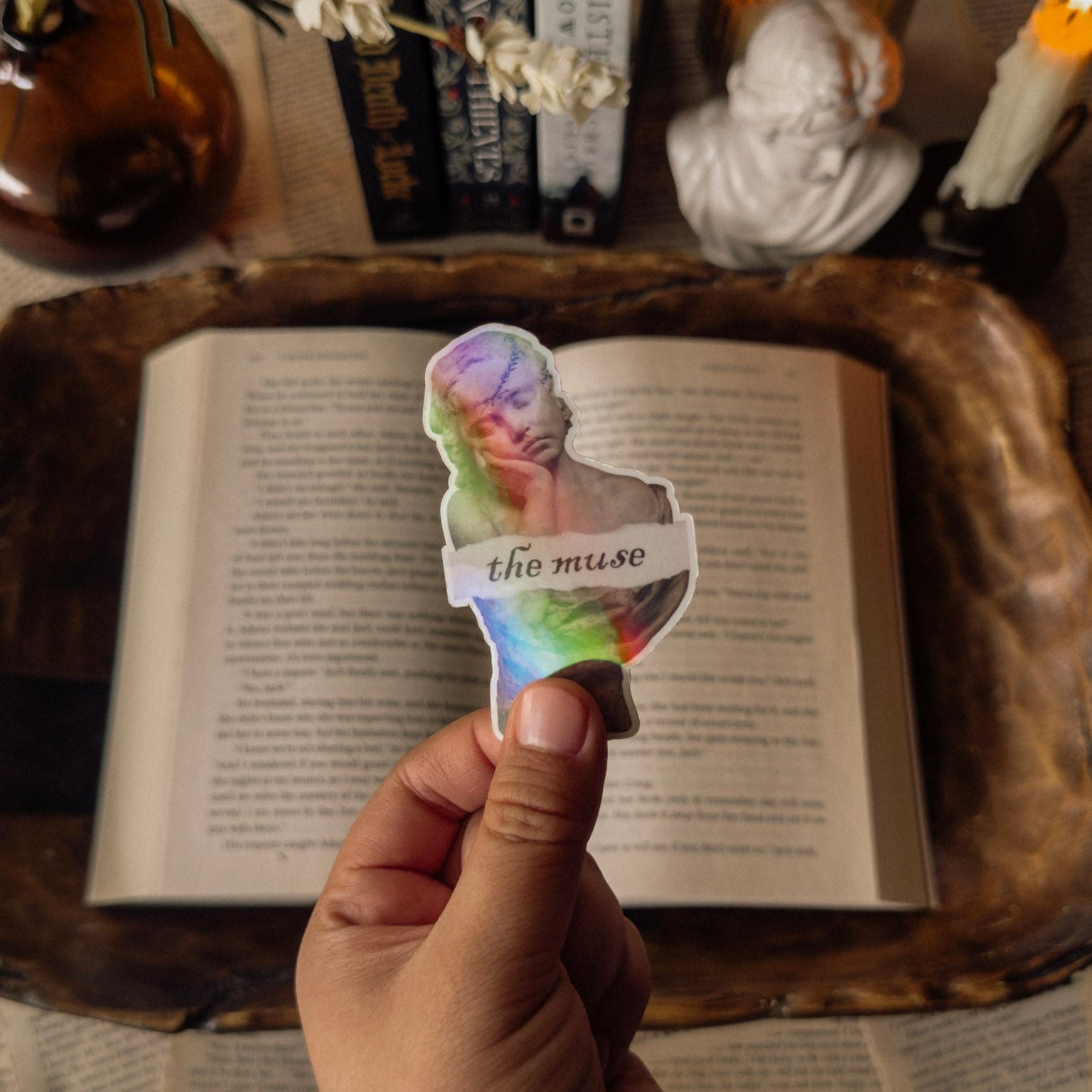 Holographic "The Muse" sticker with a prismatic rainbow finish, styled on a vintage book next to a white marble classical bust and a lit white taper candle. Perfect for a light academia aesthetic and Greek mythology lovers