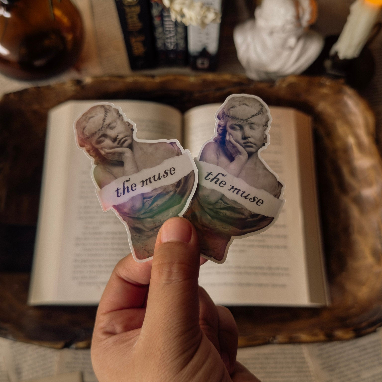 Holographic sticker featuring the text "The Muse" in serif typography, styled on an open vintage book next to a white marble classical bust and a lit white taper candle. Light academia aesthetic with a warm prismatic rainbow shimmer.