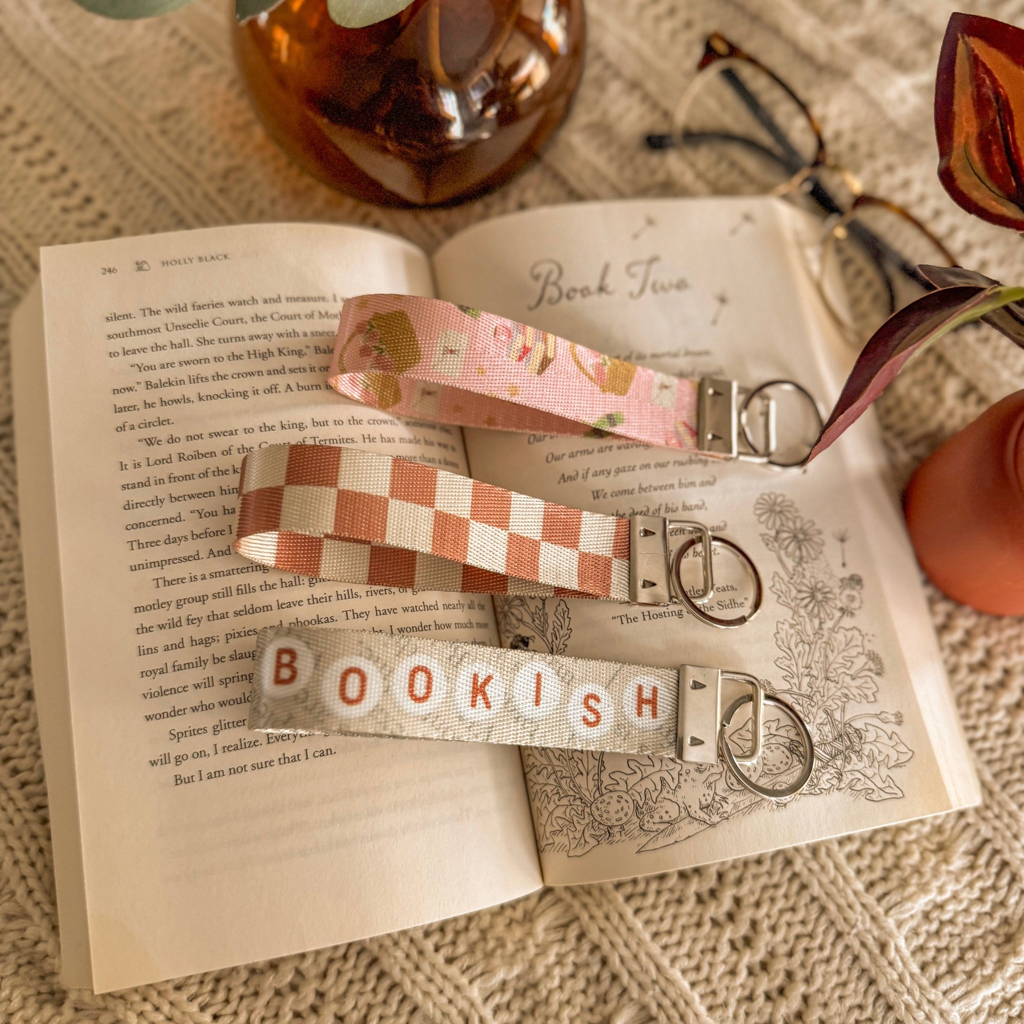 A lifestyle flat lay featuring three handmade key fobs—pink romantic floral, orange checkered, and a 'Bookish' beaded design—placed on an open vintage book. Styled with glasses and a warm drink to evoke a cozy afternoon of reading.