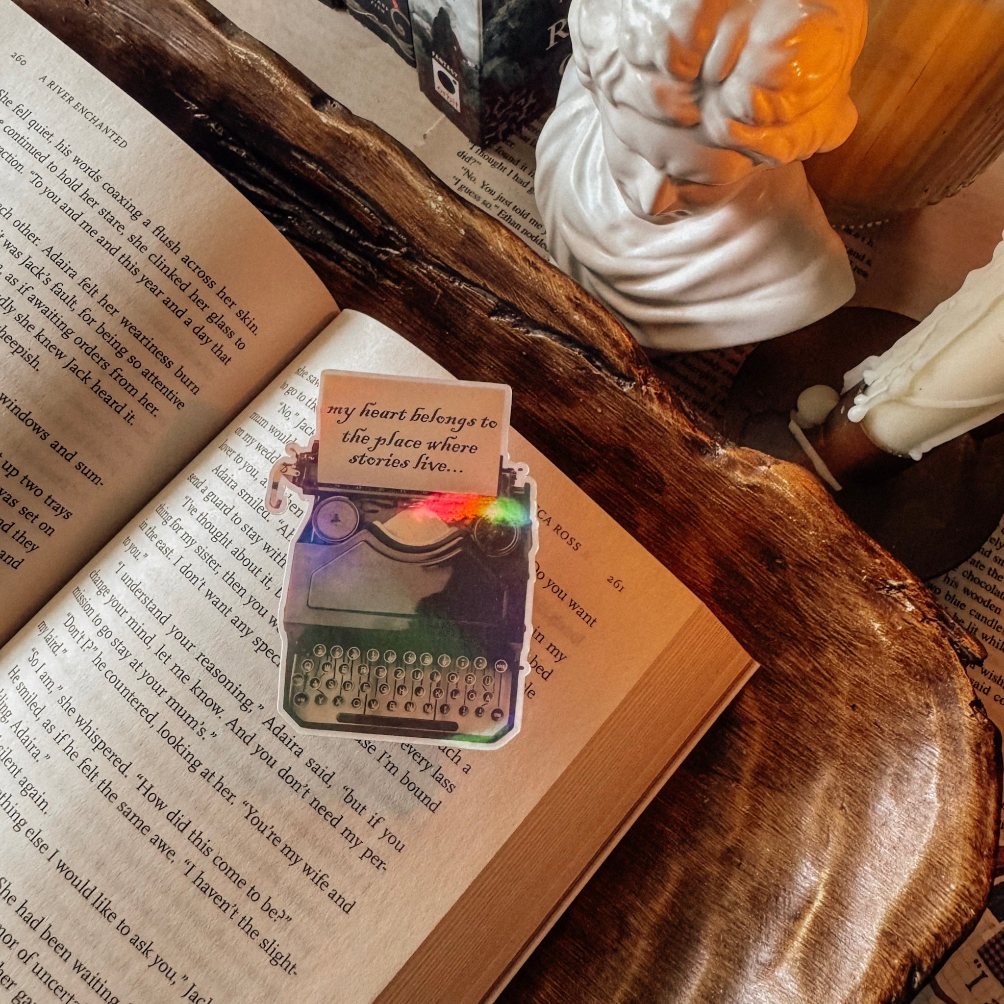 Detail shot of a holographic vintage typewriter sticker showing the prismatic rainbow effect. The sticker reads "My heart belongs to the place where stories live" and is styled in a sunlit light academia setting next to a white candle and marble bust.