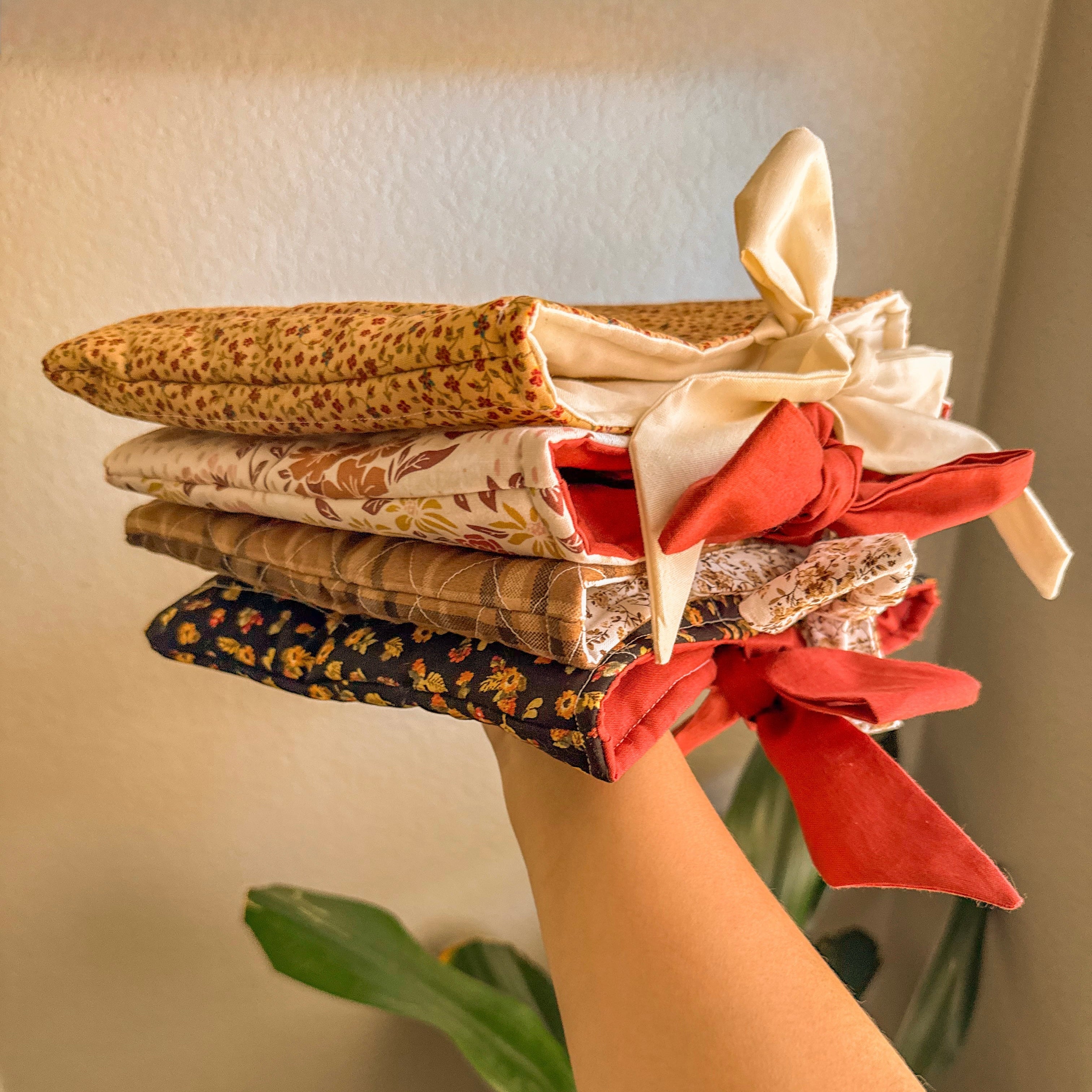 Hand holding a stack of four handmade quilted book sleeves in neutral and floral patterns with decorative fabric bows, highlighting the variety and padded protection of the Quilted Book Sleeve with Bow.