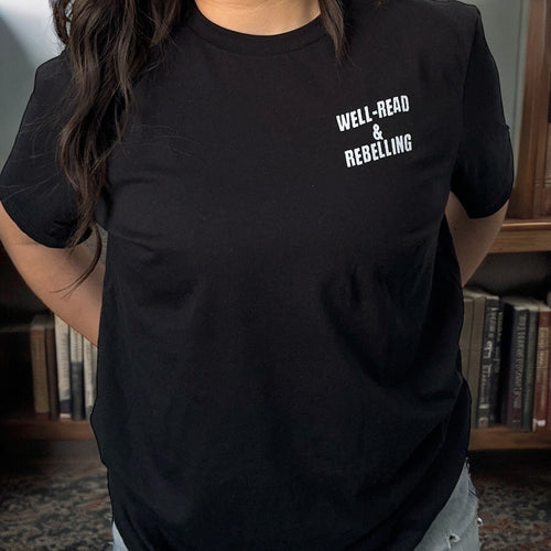 Close-up of the 'Well-Read & Rebelling' white text graphic on a black Bookish World Tour Tee by Dex Avenue, styled in front of a library bookshelf.
