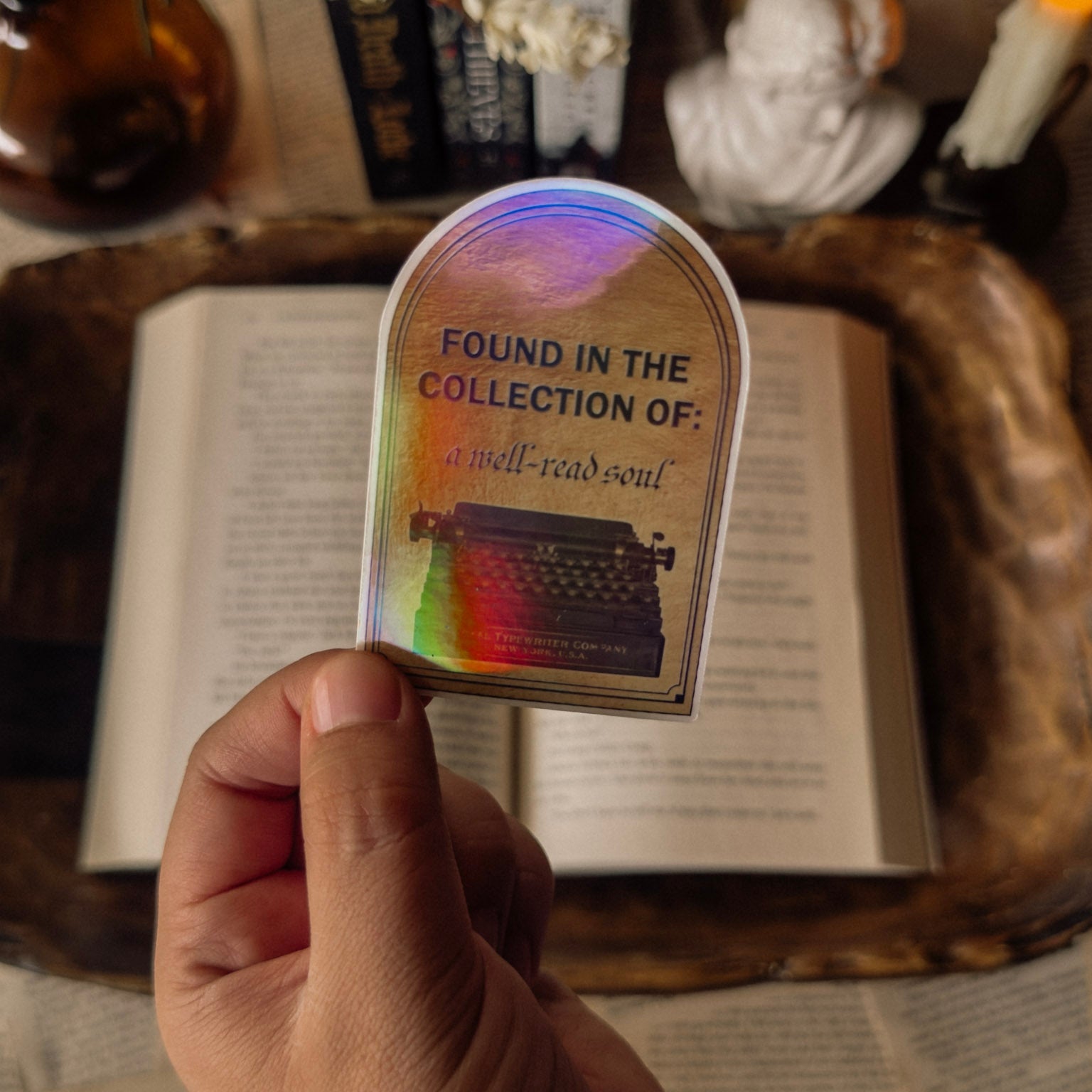 Hand holding a holographic "Found in the Collection of: a well-read soul" library-style sticker over an open book with a dark academia aesthetic and candlelight