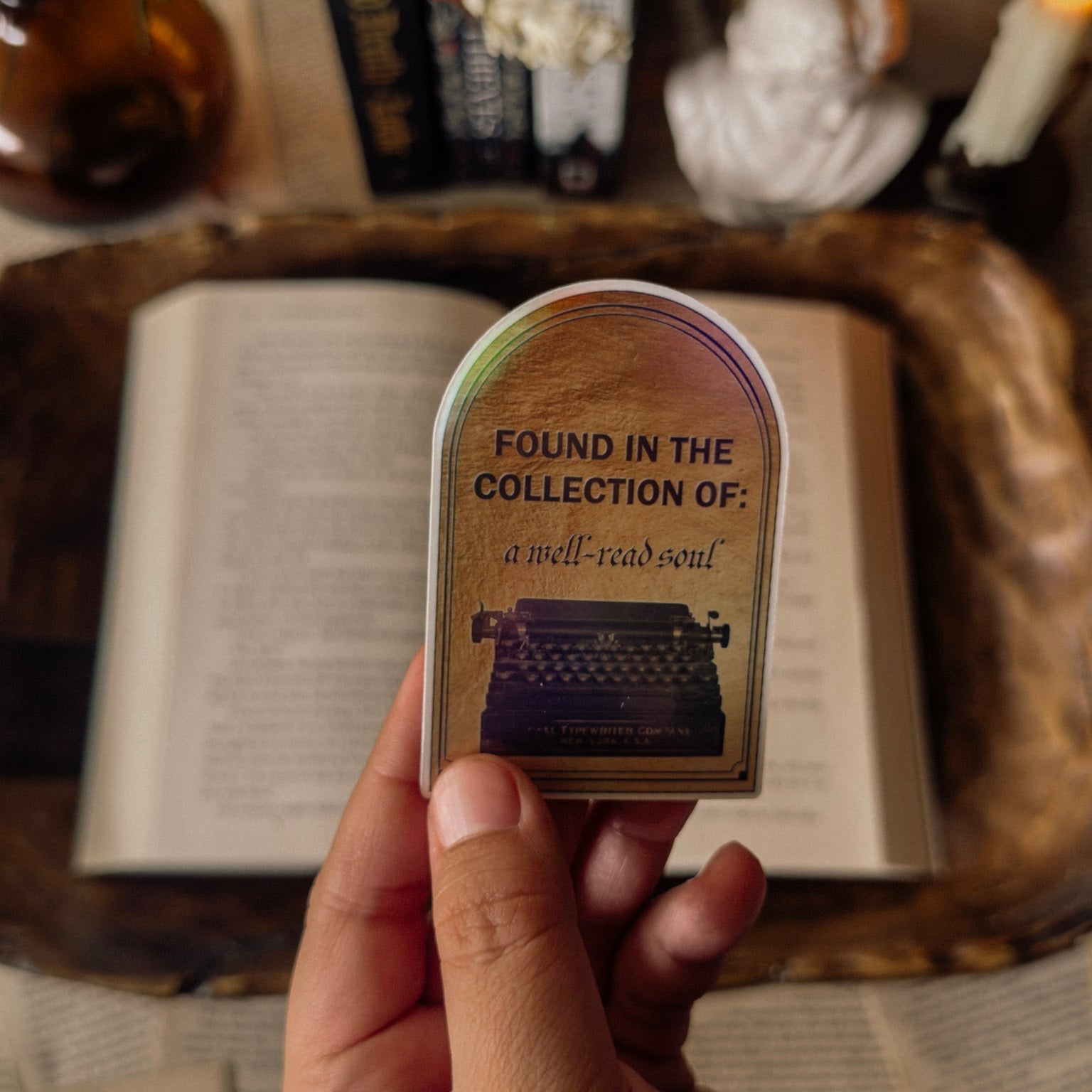 Hand holding a holographic "Found in the Collection of: a well-read soul" typewriter sticker over an open book with a dark academia library background.