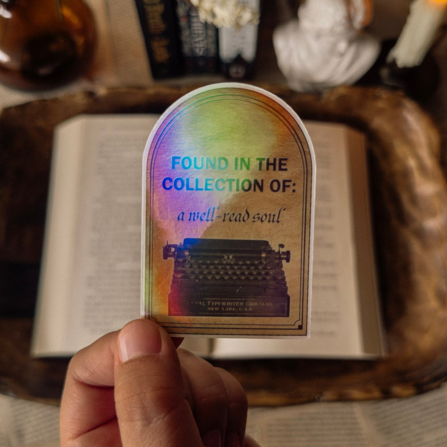 Hand holding a holographic "Found in the Collection of: a well-read soul" sticker featuring a vintage typewriter illustration, styled over an open book in a Dark Academia setting.