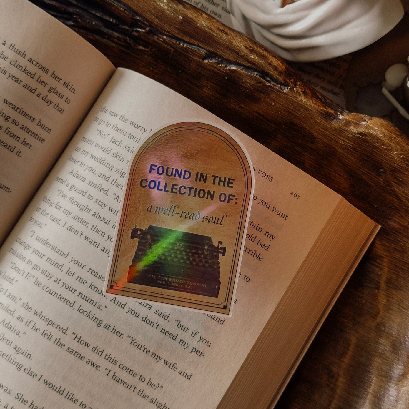 Close-up of a holographic "Found in the Collection of: a well-read soul" sticker featuring a vintage typewriter, placed on an open book in a dark academia aesthetic