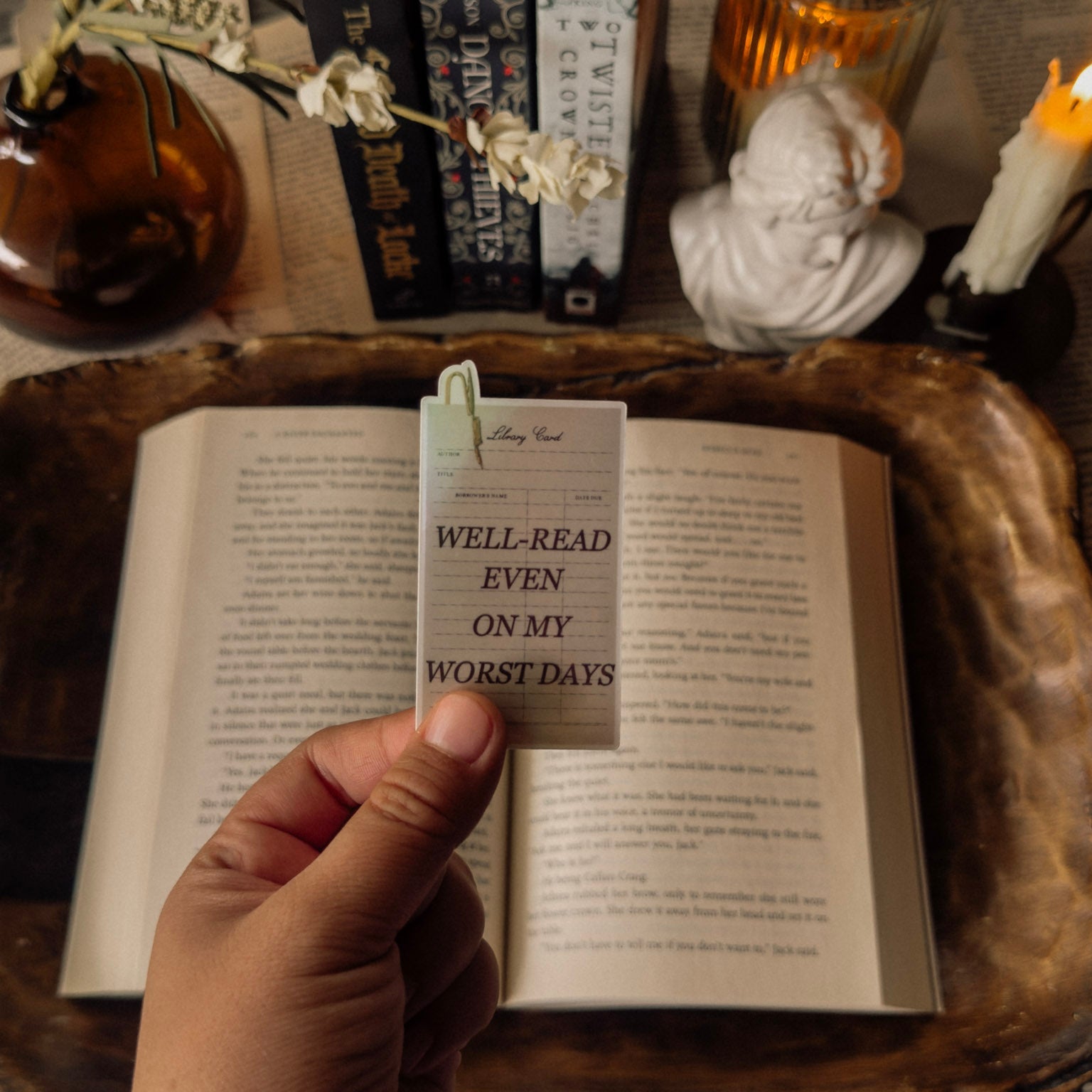 Close-up of a hand holding the "Well-Read Even on My Worst Days" holographic sticker over an open vintage book, styled with Dark Academia office decor and warm candlelight.