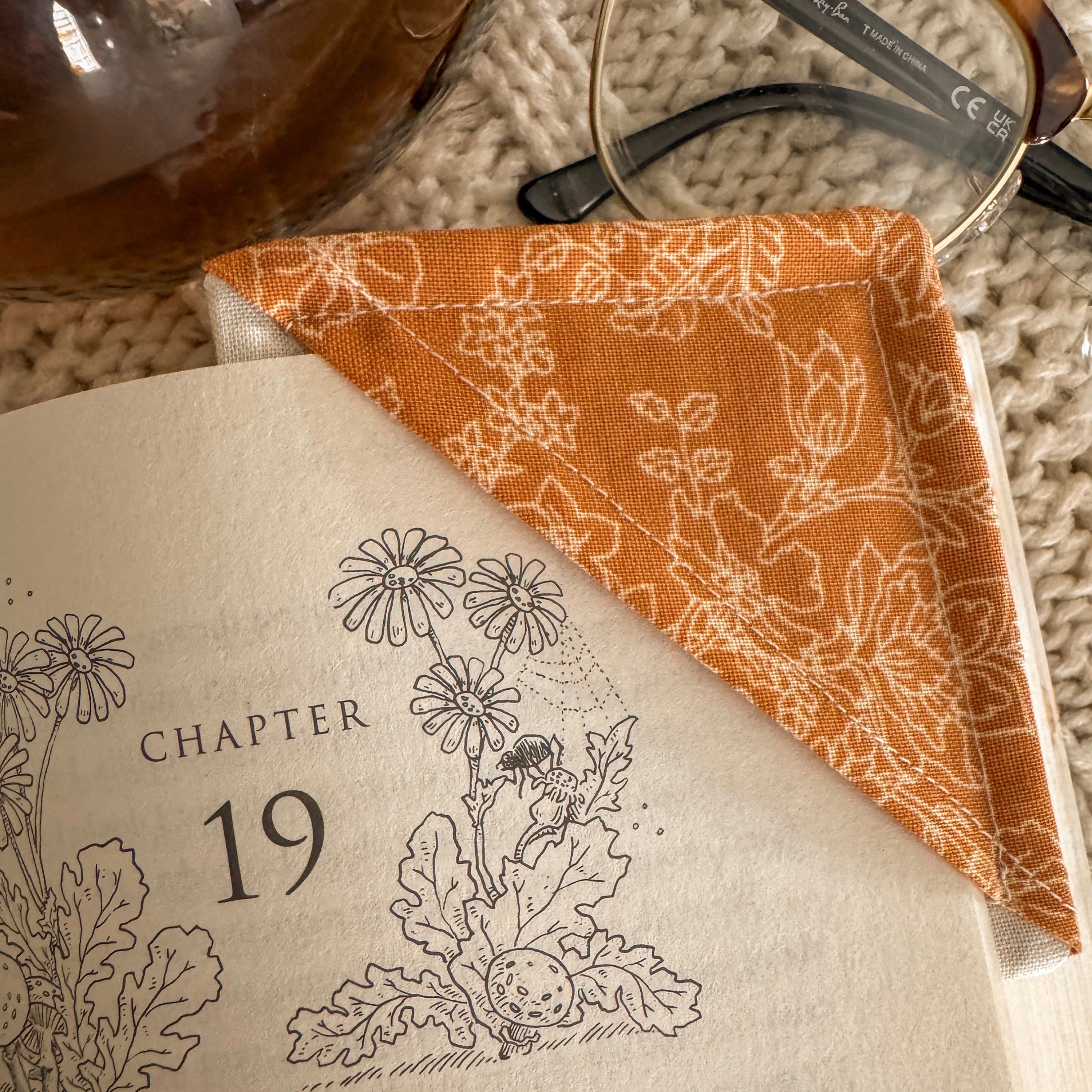 A close-up of a warm yellow floral fabric corner bookmark holding a place at Chapter 19 of a book. The intricate floral pattern and soft cotton texture are highlighted alongside vintage-style tortoiseshell glasses.